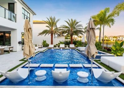 View from a pool's sun shelf with white in-water lounge chairs, looking across the water towards a modern Florida home.