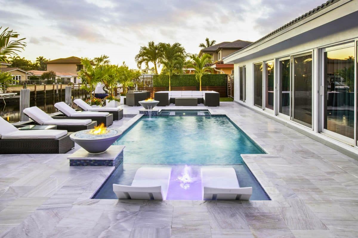 A modern waterfront pool in Fort Lauderdale with a sun shelf, raised spa, and a lit fire and water bowl at dusk.