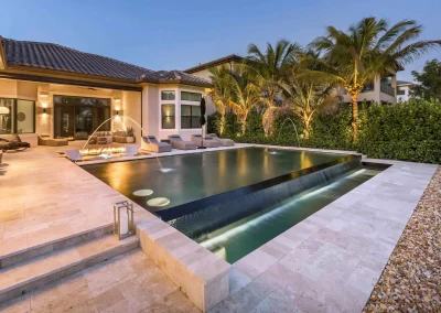 A modern geometric pool by Van Kirk Pools at dusk, with an infinity edge, three laminar deck jets, a linear fire pit, and a travertine paver patio.