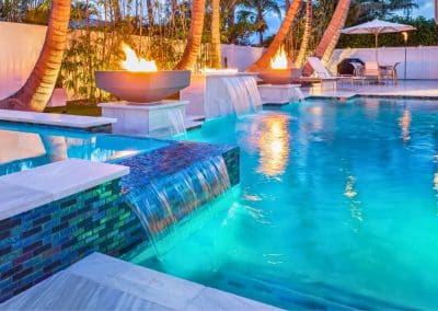A modern pool at night with a large sheer descent waterfall, two fire and water bowls, and vibrant cyan LED lighting.