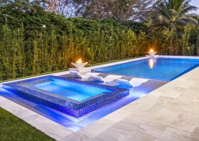 A modern Jupiter pool at dusk with a raised mosaic tile spa, a sun shelf, and two fire and water bowls.