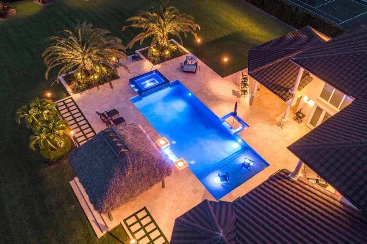 Luxury backyard pool at night with tiki hut, palm trees, built-in spa, sun shelf, and underwater LED lighting creating tropical ambiance