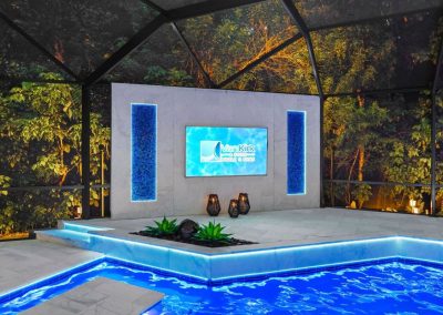 Luxury geometric pool design featuring a modern outdoor TV wall with glowing blue backlit mosaic tiles and LED pool lighting.