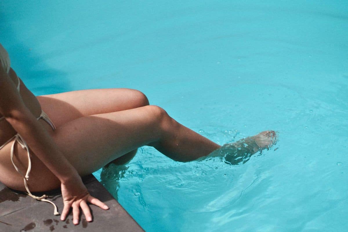 A woman in a bikini sitting on the edge of a swimming pool, dipping her feet into the cool, clear blue water.