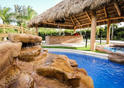 A custom rock waterfall flows into a lazy river on the "Vanilla Ice Project" pool, with a large tiki hut bar in the background, built by Van Kirk Pools.