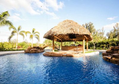 A wide view of the "Vanilla Ice Project" lagoon pool by Van Kirk Pools, showing the tiki hut bar on a central island, a rock spa, and a lazy river.
