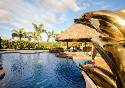 A custom gold dolphin water feature on the "Vanilla Ice Project" lagoon pool, with a tiki hut bar and rock spa in the background, built by Van Kirk Pools.