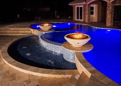 A night view of the Van Kirk Pools build for The Vanilla Ice Project, showing the multi-level pool with blue LEDs and two large fire bowls.