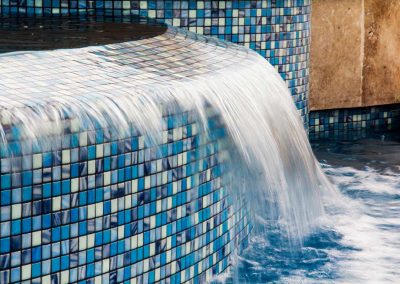 A close-up of the sheer descent waterfall from the raised spa of the "Vanilla Ice Project" pool, flowing over custom blue mosaic glass tile.