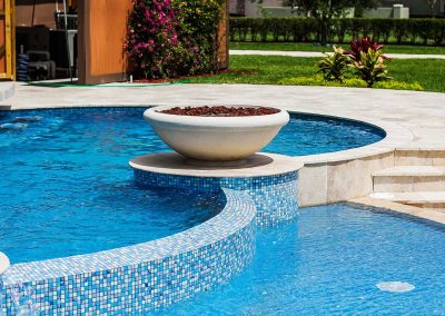 A daylight view of a large fire bowl on the raised spa of the "Vanilla Ice Project" pool, with custom blue mosaic tile, built by Van Kirk Pools.