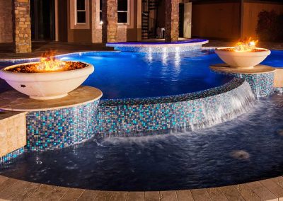 A luxury custom pool by Van Kirk Pools at night, featuring two large fire bowls, a raised spa, and a sheer descent waterfall with blue mosaic tile.