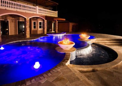 The "Vanilla Ice Project" pool by Van Kirk Pools at night, with two large fire bowls, a sheer descent waterfall, and bright blue LED lights.