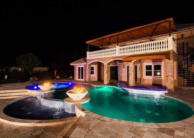 A wide night shot of the Van Kirk pool from "The Vanilla Ice Project" with two large fire bowls, a swim-up bar, a raised spa, and LED lights.