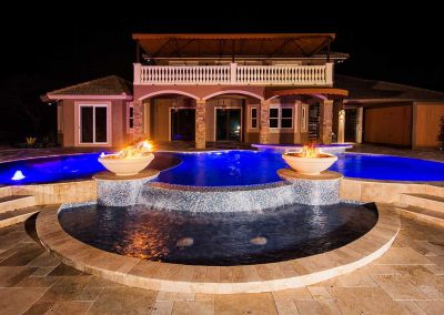 The "Vanilla Ice Project" pool by Van Kirk Pools at night, with two fire bowls, a mosaic tile waterfall, a raised spa, and blue LED lighting.