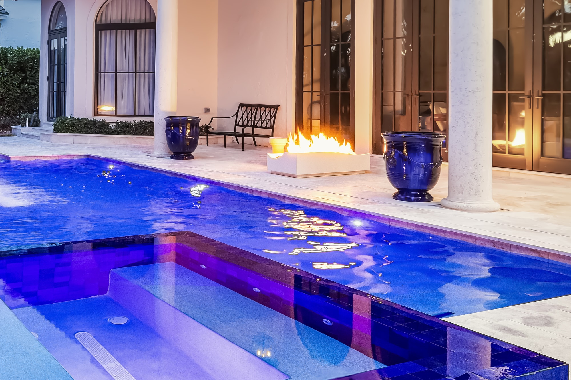Luxury patio with a glowing pool and fire pit at sunset, surrounded by modern outdoor seating and palm trees.