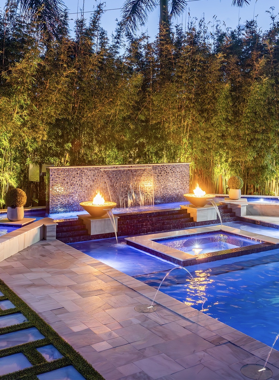 Colorful luxury pool with LED lights and fire feature, set against a tropical garden backdrop at sunset.