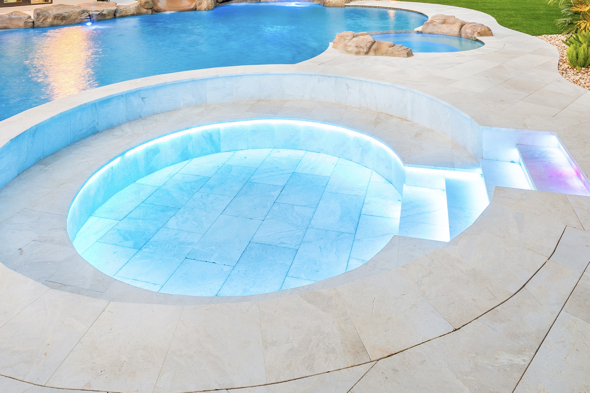 Luxury poolside view at night with illuminated water, lush palm trees, and decorative rock waterfall.