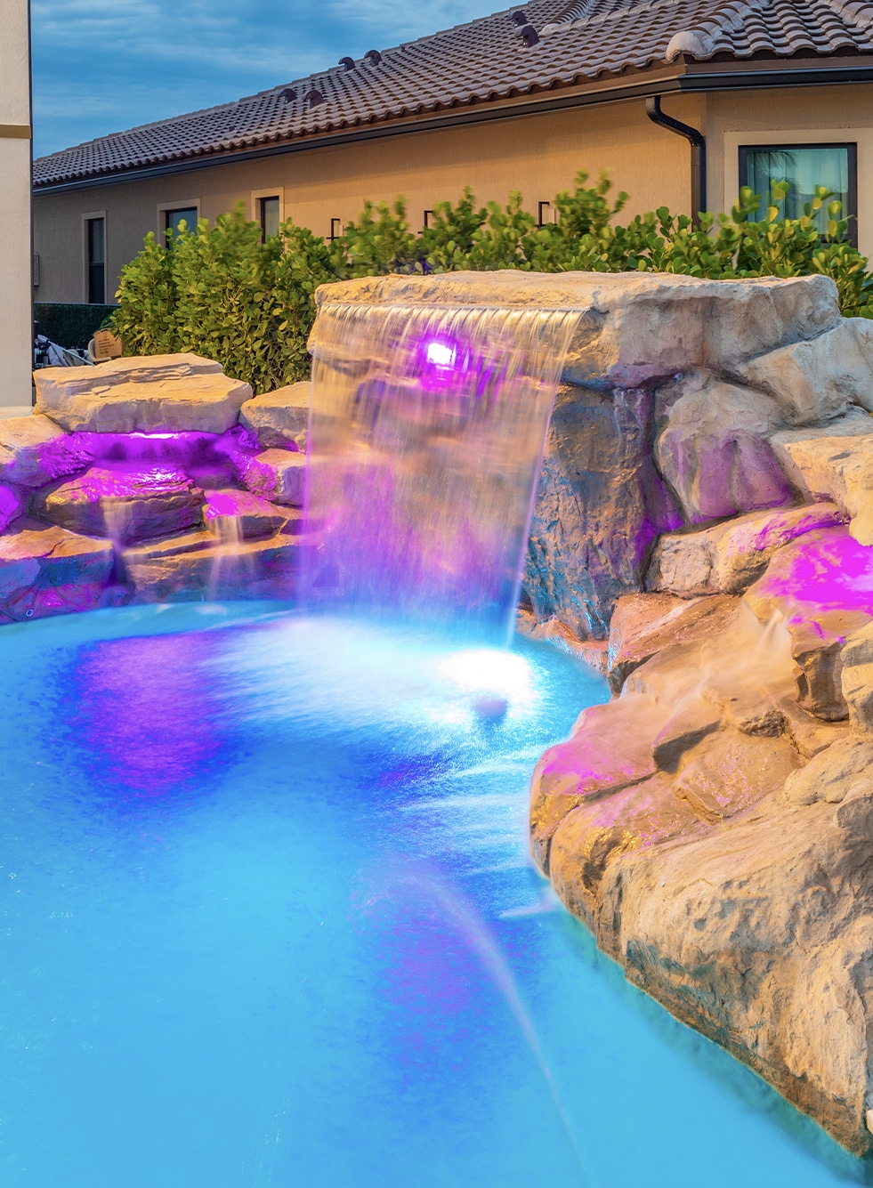 Luxurious backyard pool with waterfalls, fire bowls, and vibrant lighting under palm trees at dusk.