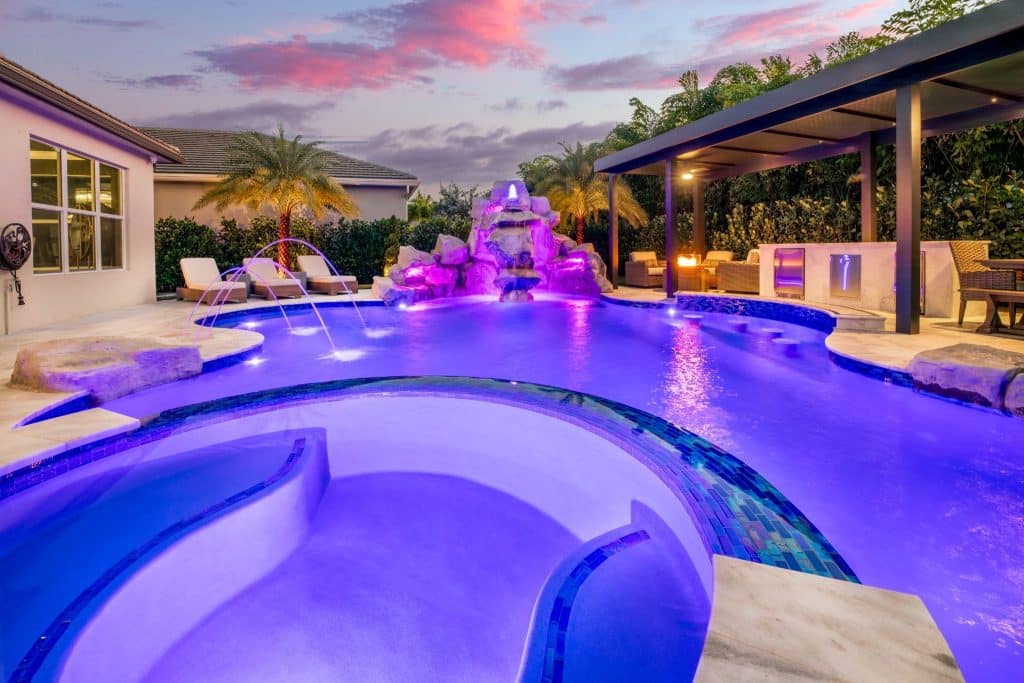 Backyard swimming pool at dusk with purple lights, a rock waterfall, palm trees, and lounge chairs under a covered patio.