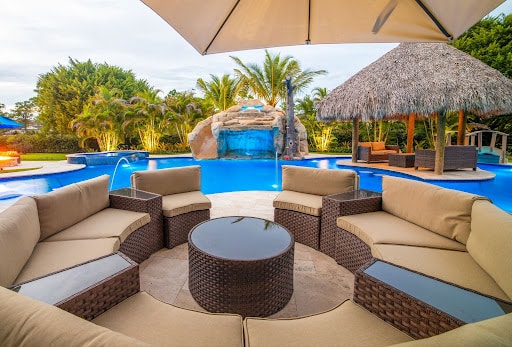 Circular wicker lounge with beige cushions around a round table under a large umbrella, by a pool with a thatched cabana and palm trees in the background.
