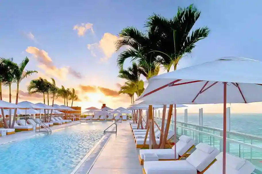 1-Hotel-Rooftop-Pool-at-Sunset-Miami-Beach_11zon - Van Kirk & Sons Pools & Spas Sunset view of a tropical resort pool deck with palm trees, white lounge chairs, and umbrellas overlooking the ocean.